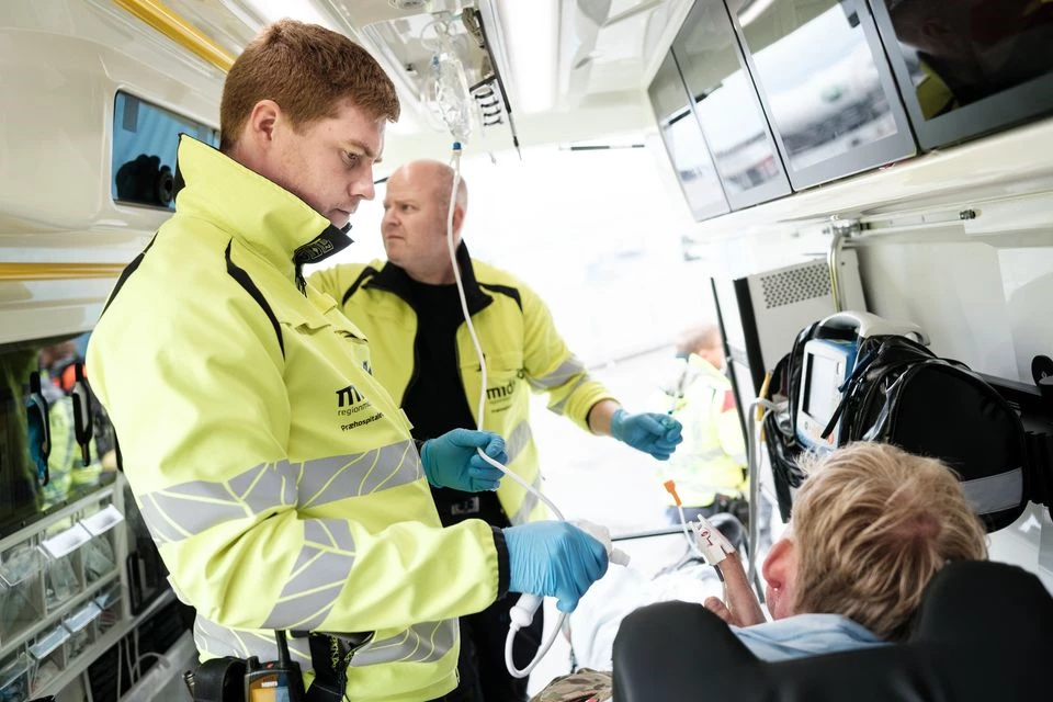 To reddere står indeni en ambulance og kigger på en patient, der ligger på en båre. 